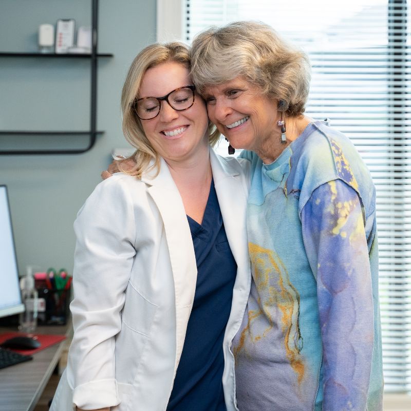 Dr. Grome, in glasses and a white coat, smiling as she shares a hug with an older patient